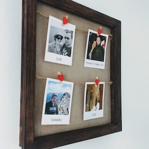 May include: A brown wooden picture frame with a burlap background and four photos held in place with red heart-shaped clothespins. The photos are of a couple in different locations, including Crete, Snowdonia, and a wedding in Poland. The text below each photo identifies the location.