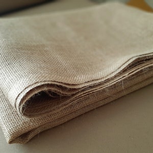 May include: A folded piece of natural brown burlap fabric. The fabric is woven with a loose, open weave and has a rough texture.