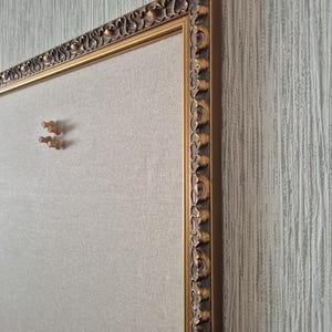 Op de afbeelding: Een rechthoekig prikbord met een lichtbeige stoffen oppervlak, omlijst door een sierlijke, antiek goudkleurige rand met een herhalend krulpatroon. Twee kleine, ronde punaises van lichtgekleurd hout zijn bevestigd op het stoffen oppervlak.