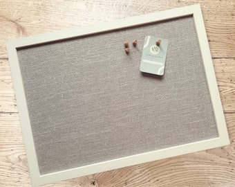 May include: A rectangular bulletin board with a light gray fabric surface and a white wooden frame. Three wooden push pins and a business card are attached to the board. The business card has the text "A Bethany Handmade".