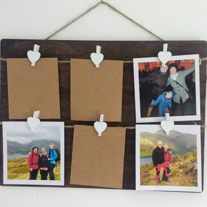 May include: A wooden photo display with six spaces for photos. Each space has a white heart-shaped clip to hold the photos in place. The photos are of people in various settings, including a group of people hiking in the mountains, a couple standing on a hill overlooking a lake, and a group of people at a concert.