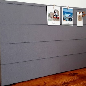 May include: A gray fabric bulletin board with horizontal lines, featuring three photos pinned with clothespins. The photos show a red building, a beach scene, and a brown tag with the text "Greetings".