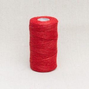 May include: A spool of red twine. The twine is tightly wound around a central core, creating a cylindrical shape. The colour is a vibrant, solid red, and the texture appears slightly rough. The background is a neutral off-white.