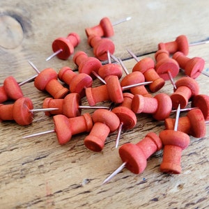 May include: A pile of red wooden push pins with silver metal points. The push pins are scattered on a wooden surface.