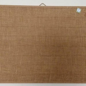 May include: A brown cork board with a woven texture. The board has a metal hanger at the top and is mounted on a white wall. There are two small white tags pinned to the board.