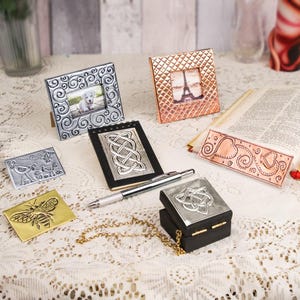 May include: An assortment of decorative metal items, including picture frames, a notepad, and a small box, are arranged on a lace tablecloth. The frames feature intricate designs and hold images. A pen and a gold chain are also present.