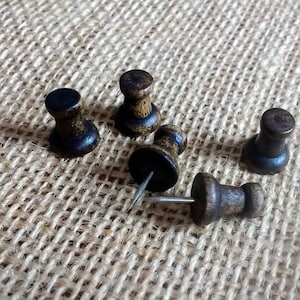 May include: A set of six wooden push pins with black heads and silver metal pins. The push pins are arranged on a brown hessian surface.