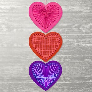 May include: Three hanging heart-shaped decorations in pink, red, and purple. Each heart has a unique stitched design with contrasting thread colours. The decorations are suspended by thin strings against a grey wooden background.