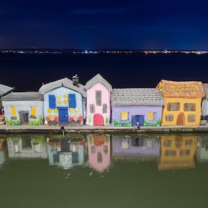 May include: A row of five miniature model houses in gray, light blue, pink, lavender, and yellow, each with distinct doors, windows, and roof textures, positioned along a dark water edge reflecting the colorful structures at night.