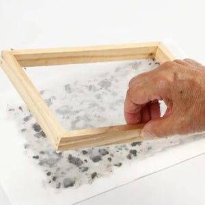 May include: A wooden frame with a mesh screen is held over a sheet of paper with a speckled design. The frame is light brown, and the paper has a white background with gray and black spots. The image suggests a craft project or handmade paper.