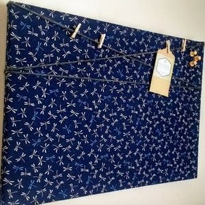 May include: A blue fabric bulletin board with a dragonfly pattern. The board has string and wooden clothespins for hanging items. There is a small brown paper tag with the text "Handmade with Love" on the board.