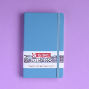 May include: A blue sketchbook with a white elastic band closure. The cover has a label with the text "art creation SKETCH BOOK 80 SHEETS/VEL/FEUILLES/BOGEN/HOJAS/LENZUOLA 1521CH/5583-3400/M/90LBS ACID FREE".