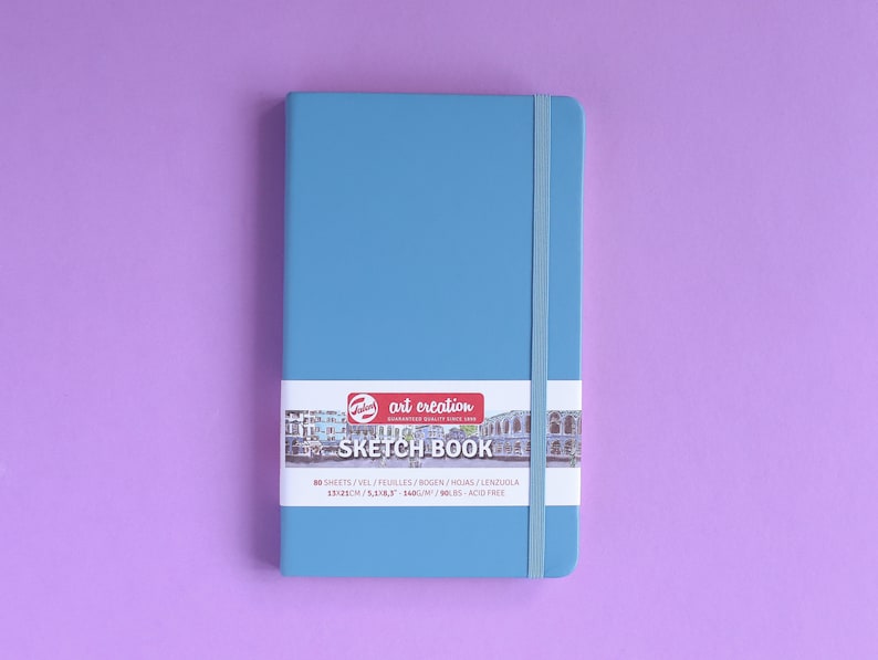 May include: A blue sketchbook with a white elastic band closure. The cover has a label with the text "art creation SKETCH BOOK 80 SHEETS/VEL/FEUILLES/BOGEN/HOJAS/LENZUOLA 1521CH/5583-3400/M/90LBS ACID FREE".