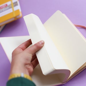 May include: A pink notebook with a white ribbon bookmark and a blank page open. The notebook is on a purple surface.