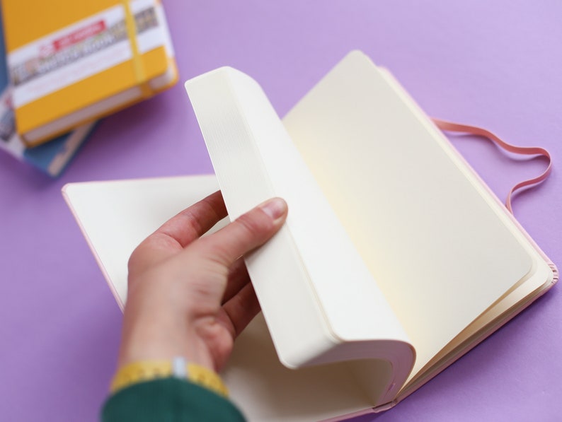 May include: A pink notebook with a white ribbon bookmark and a blank page open. The notebook is on a purple surface.