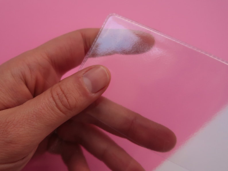 May include: A clear plastic sheet protector, held in a hand, with a pink background.