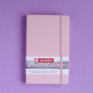 May include: A pink sketchbook with a black elastic band closure. The cover has a sticker with the text "art creation" and "SKETCH BOOK" in white lettering. The sticker also includes the text "80 SHEETS/VIEL/FEUILLES/BOGEN/HOJAS/LENZUOLA 13.323CM/5.243" in black lettering.