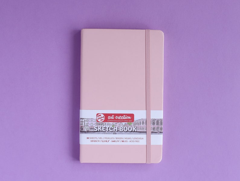 May include: A pink sketchbook with a black elastic band closure. The cover has a sticker with the text "art creation" and "SKETCH BOOK" in white lettering. The sticker also includes the text "80 SHEETS/VIEL/FEUILLES/BOGEN/HOJAS/LENZUOLA 13.323CM/5.243" in black lettering.