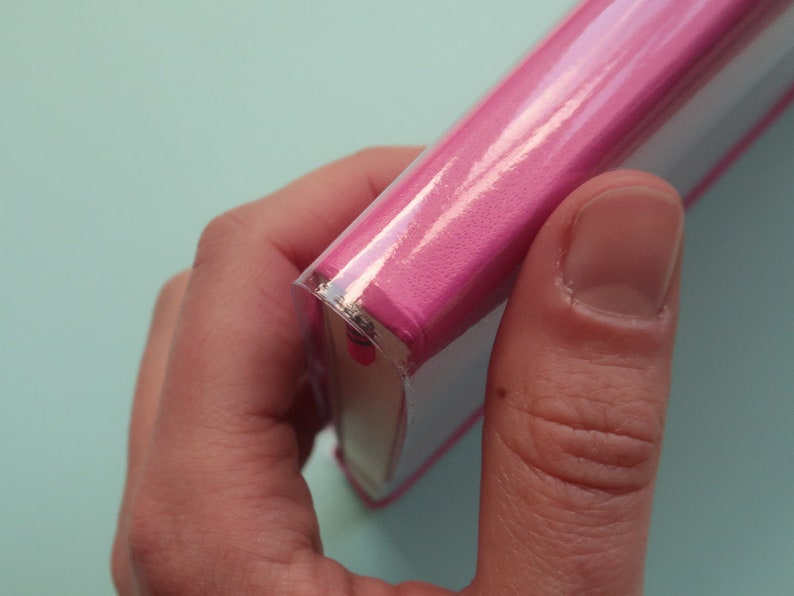 May include: A pink book with a clear plastic cover. The book is being held by a hand.