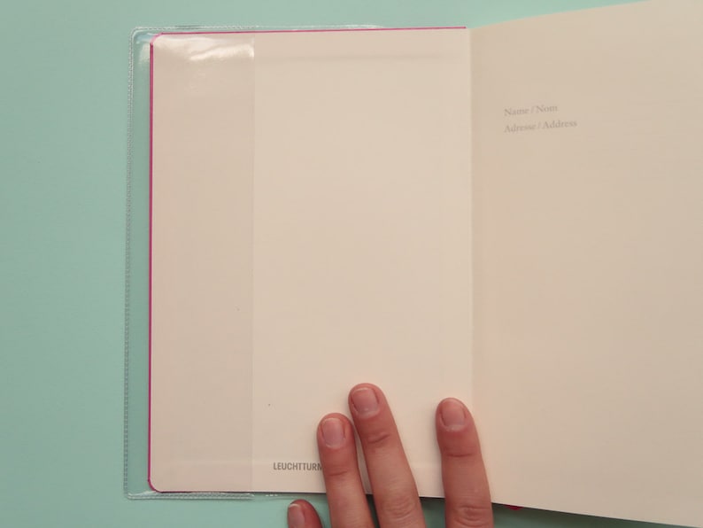 May include: An open notebook with a pink and clear plastic cover. The notebook has a blank page with the words "Name/Nom" and "Address/Adresse" written in black ink.