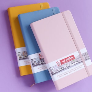 May include: Three sketchbooks in different colors: yellow, blue, and pink. Each sketchbook has a black elastic band closure and a label with the text "art creation sketchbook" and details about the paper.