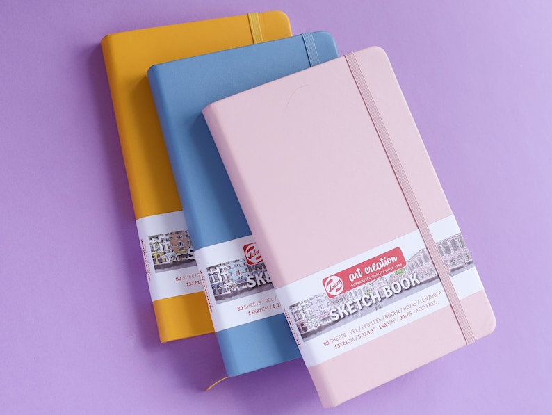 May include: Three sketchbooks in different colors: yellow, blue, and pink. Each sketchbook has a black elastic band closure and a label with the text "art creation sketchbook" and details about the paper.
