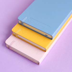 May include: Three notebooks stacked on top of each other. The top notebook is light blue with the text "The Daily Routine" on the cover. The middle notebook is yellow with the text "The Daily Routine" on the cover. The bottom notebook is pink with the text "The Daily Routine" on the cover.