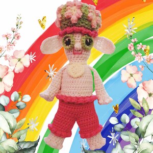May include: A handmade crochet doll with a whimsical design. The doll features a pink body, red pants, and a hat with pink and green accents. It has large, expressive eyes and a smiling face. The background includes a rainbow and floral elements.