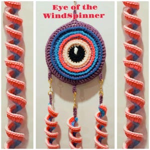 Puede incluir: Un tapiz de pared de ganchillo hecho a mano con el texto "Eye of the WindSpinner". El diseño circular presenta anillos concéntricos de colores azul, coral y crema, que se asemejan a un ojo. Se adjuntan detalles en espiral decorativos en rojo, blanco y azul.