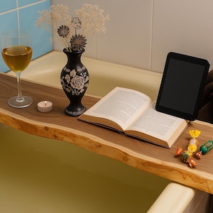 May include: A wooden bathtub tray with a live edge, holding a glass of wine, a small black vase with flowers, a lit candle, an open book, a tablet, and wrapped candies. The tray is made of light brown wood.