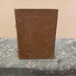 Puede incluir: Un libro encuadernado en cuero marrón con un diseño decorativo en la portada. El título en la portada dice "Westphalia's New German Grammar".