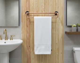 Puede incluir: Un toallero de doble barra construido con tuberías de metal color cobre con soportes de pared circulares negros. Una toalla rectangular blanca cuelga sobre la barra superior del toallero, montado en un panel de pared de madera clara.
