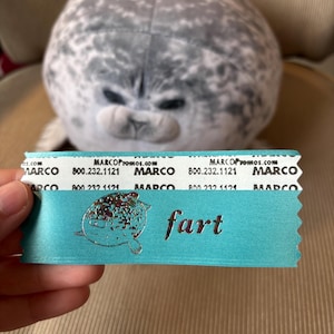 May include: A plush gray seal toy with a light blue ribbon that says "fart" in silver lettering. The ribbon also has the text "MARCOPromos.com" and a phone number.