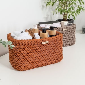 May include: Two crocheted storage baskets, one brown and one beige, on a white countertop. The brown basket contains toiletries, including bottles of shampoo and conditioner, and a white hand towel. The beige basket is empty.