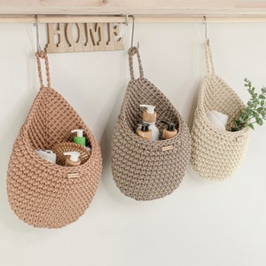Puede incluir: Tres cestas de almacenamiento de crochet colgantes con asas de cuerda. Las cestas son beige, marrón y blanco. La cesta beige contiene artículos de tocador, la cesta marrón contiene artículos de tocador y la cesta blanca contiene una toalla y una planta.