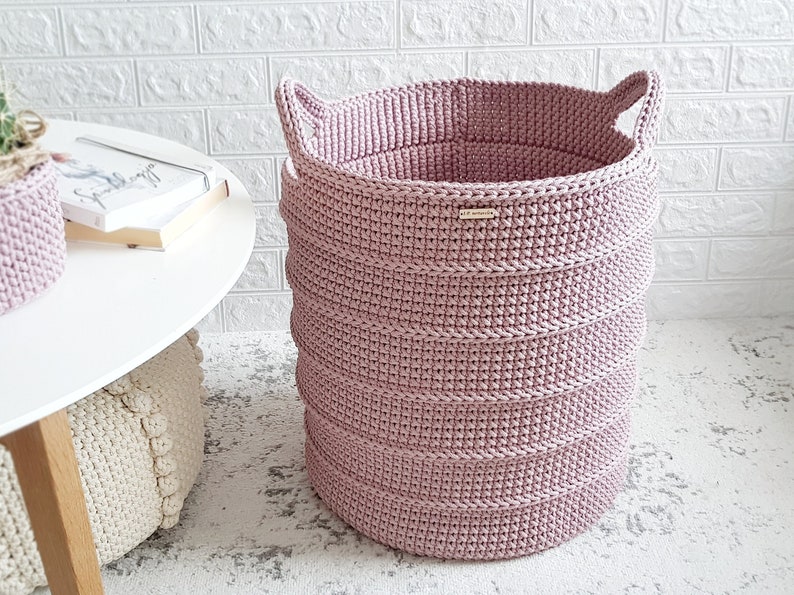 Puede incluir: Una cesta de almacenamiento grande de color rosa hecha a crochet con dos asas. La cesta est&aacute; hecha de un hilo grueso y texturizado y tiene una parte superior ancha y abierta.