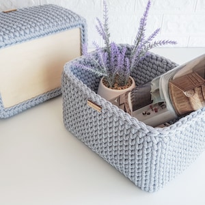 May include: Two gray crocheted storage baskets. One basket is empty and the other basket contains a potted plant and a magazine.