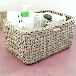 May include: A beige, rectangular, crocheted storage basket filled with white towels, skincare products, and a jade roller. The basket has a small wooden tag and sits on a pink surface. The basket is made of thick yarn.