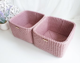 May include: Two pink crocheted storage baskets with wooden handles. The baskets are made of a soft, textured yarn and are perfect for organizing small items.