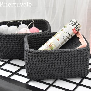 Puede incluir: Dos cestas de almacenamiento de crochet grises. Una cesta está llena de bolas de algodón blancas y rosas. La otra cesta contiene una revista y un teléfono. Un carrete de hilo rosa está en el fondo.