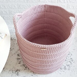Puede incluir: Una cesta de almacenamiento de crochet rosa con dos asas. La cesta est&aacute; hecha de hilo grueso y tiene una textura tejida.