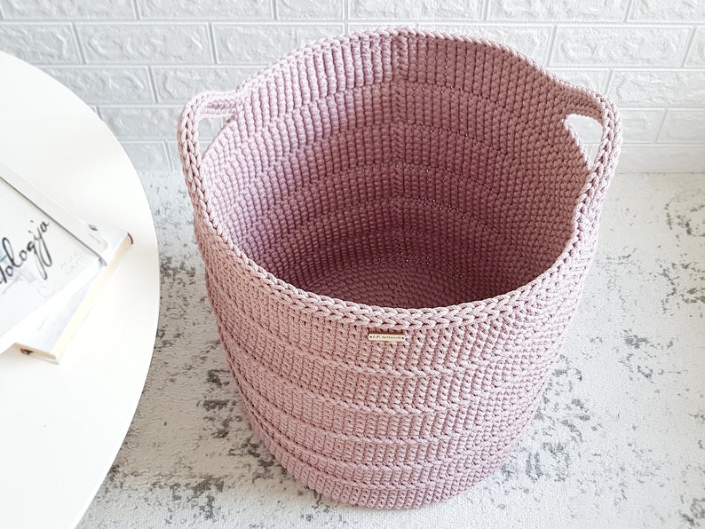 Puede incluir: Una cesta de almacenamiento de crochet rosa con dos asas. La cesta est&aacute; hecha de hilo grueso y tiene una textura tejida.