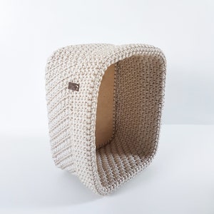May include: A beige crocheted storage basket with a rectangular shape. The basket is made of a thick, textured yarn and has a slightly open top.