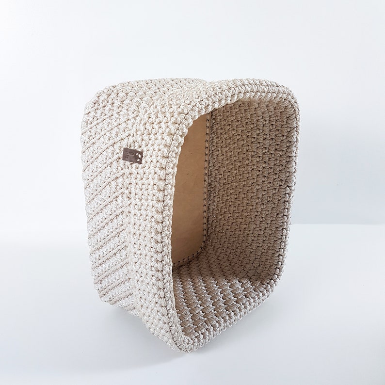 May include: A beige crocheted storage basket with a rectangular shape. The basket is made of a thick, textured yarn and has a slightly open top.