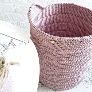 Puede incluir: Una cesta de almacenamiento de crochet grande de color rosa con dos asas. La cesta est&aacute; hecha de hilo grueso y tiene una textura tejida.
