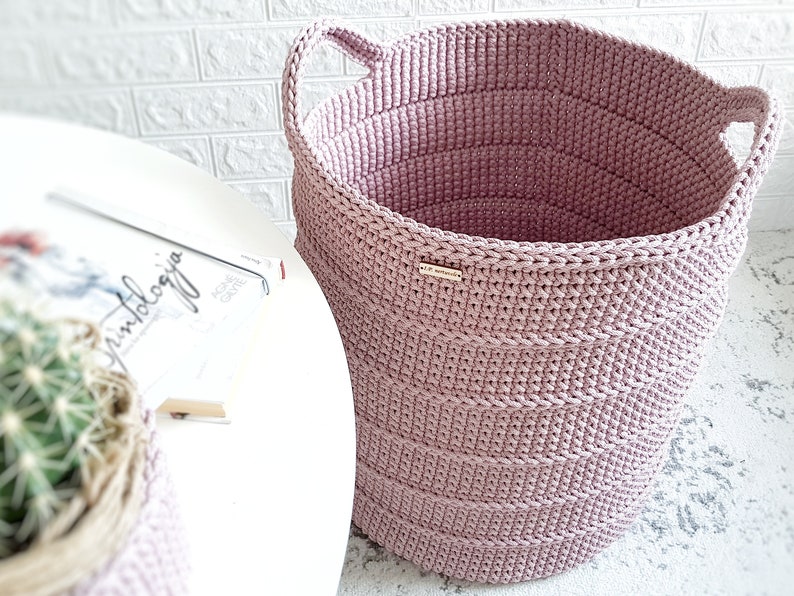 Puede incluir: Una cesta de almacenamiento de crochet grande de color rosa con dos asas. La cesta est&aacute; hecha de hilo grueso y tiene una textura tejida.