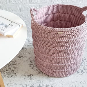 Puede incluir: Una cesta de almacenamiento de crochet rosa con dos asas. La cesta est&aacute; sobre una alfombra blanca.