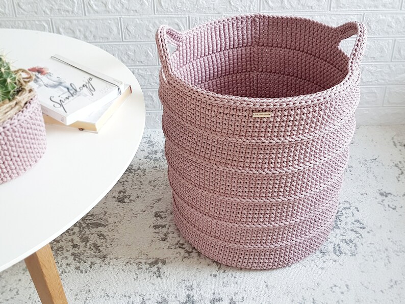 Puede incluir: Una cesta de almacenamiento de crochet rosa con dos asas. La cesta est&aacute; sobre una alfombra blanca.