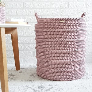 Puede incluir: Una cesta de lavander&iacute;a de crochet rosa con dos asas. La cesta est&aacute; sentada en una alfombra blanca frente a una pared de ladrillo blanco.