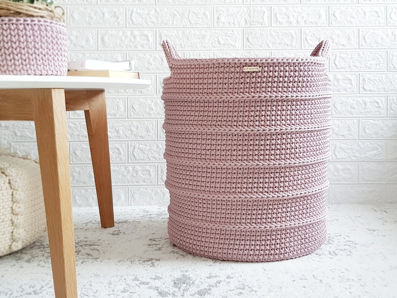 Puede incluir: Una cesta de lavander&iacute;a de crochet rosa con dos asas. La cesta est&aacute; sentada en una alfombra blanca frente a una pared de ladrillo blanco.